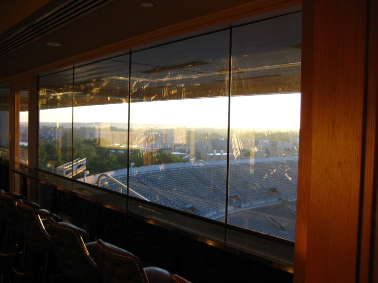 Early morning sunlight revealing widespread scratches across 8,500 sq ft of tempered glass at Notre Dame Stadium—view looking out toward stadium seating areas